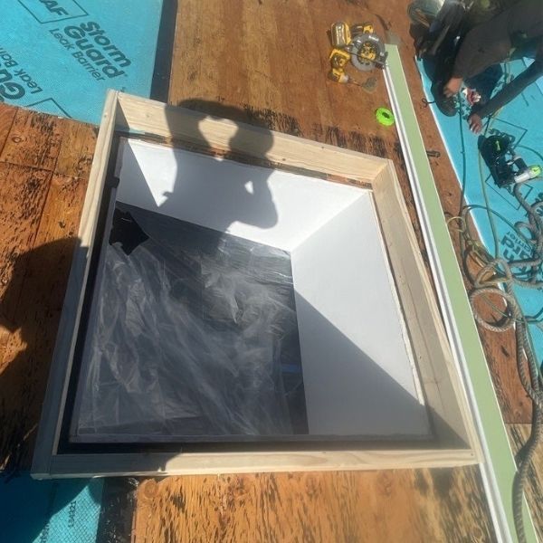 A square wooden frame for a skylight installation on a roof covered in blue underlayment, with construction tools nearby.