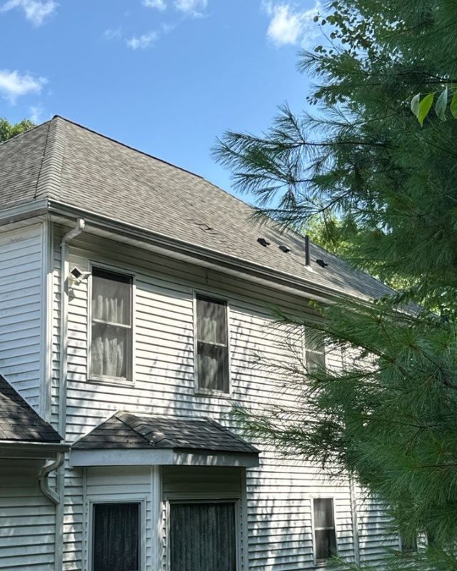 Two-story white house with gray shingled roof, windows, and trees on a sunny day.