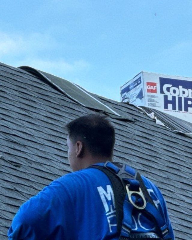 Roofer wearing safety harness on a roof, working near a damaged area. A box of roofing materials is visible.