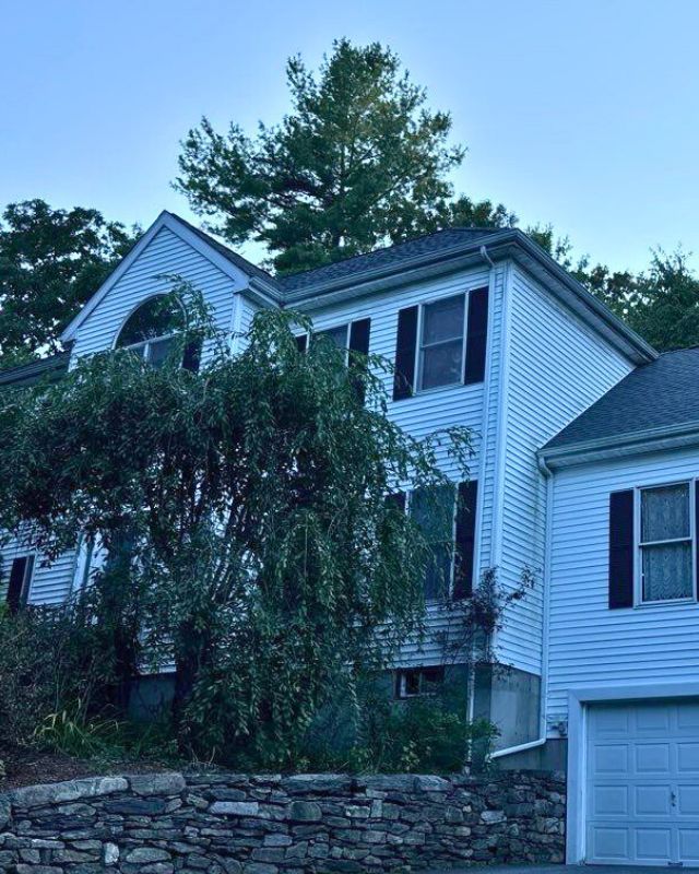 Two-story white house with black shutters, visible roof, and attached garage. A tree partially obscures the house.