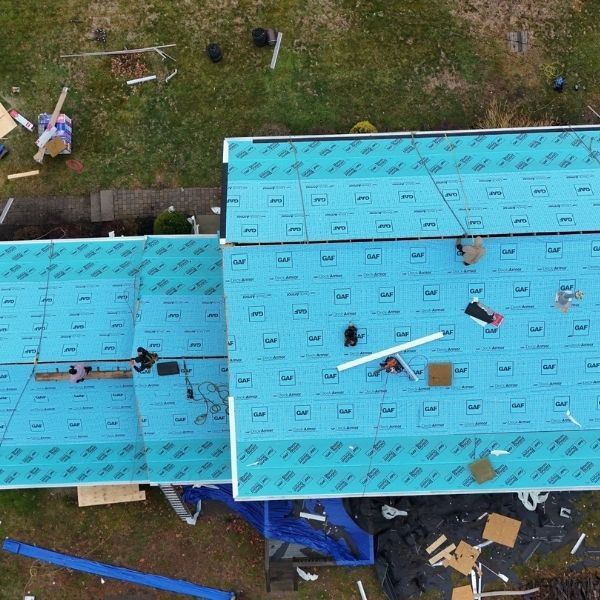 Aerial view of workers installing blue roofing underlayment on a multi-section house roof.