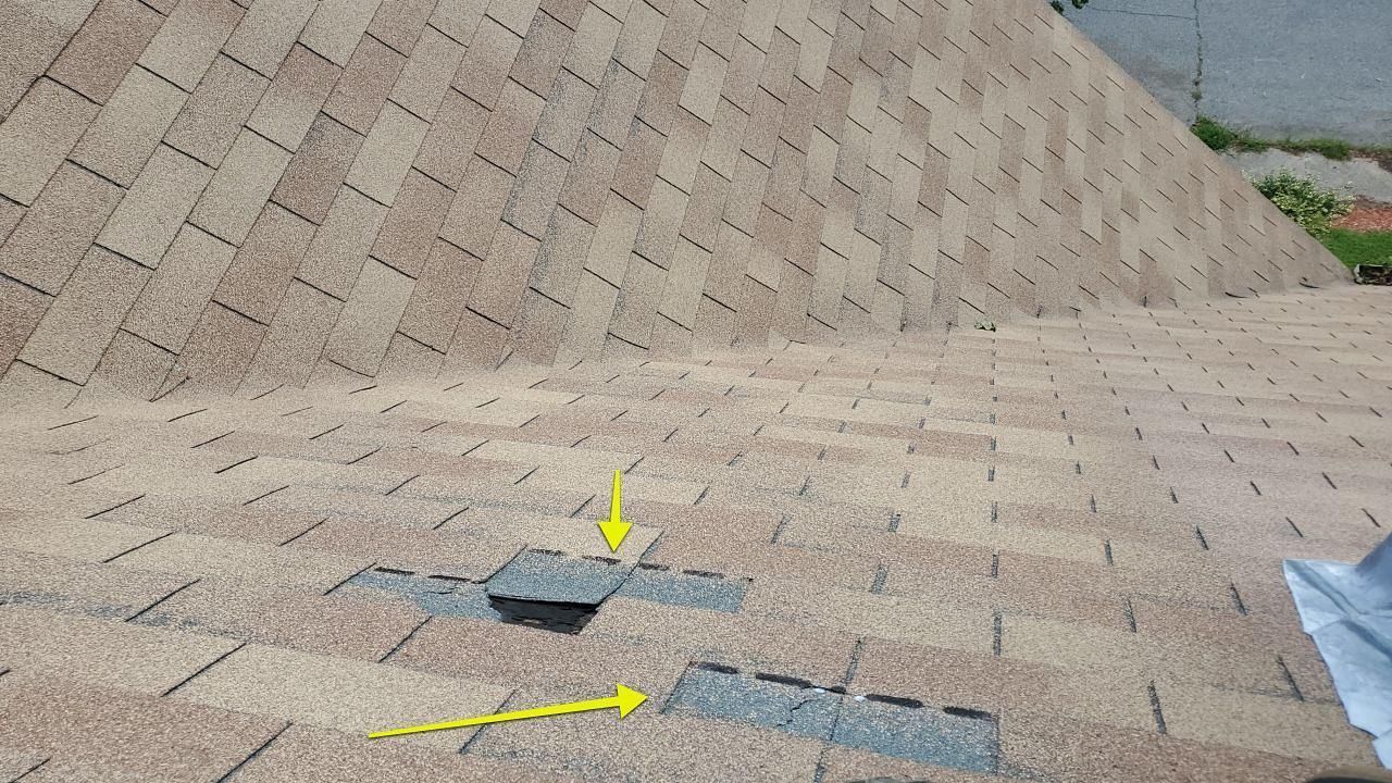 Damaged asphalt shingles on a roof, with two areas where shingles are missing and indicated by yellow arrows.