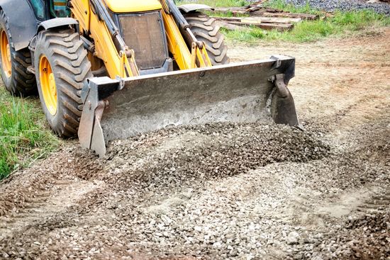 Yellow backhoe spreading gravel on a dirt road construction site