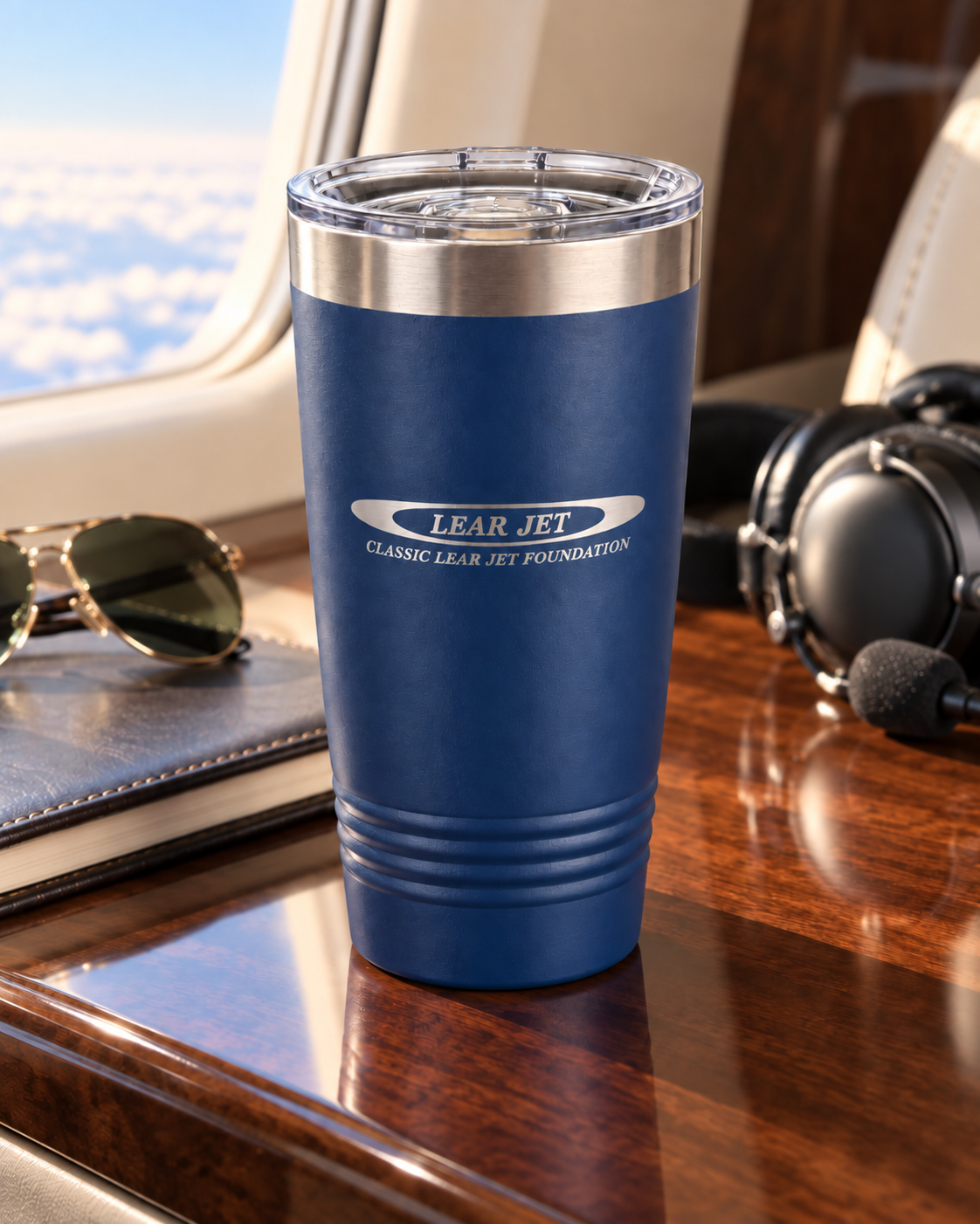 Blue 20 oz tumbler on a wooden airplane tray table with sunglasses, headphones, and a notepad nearby