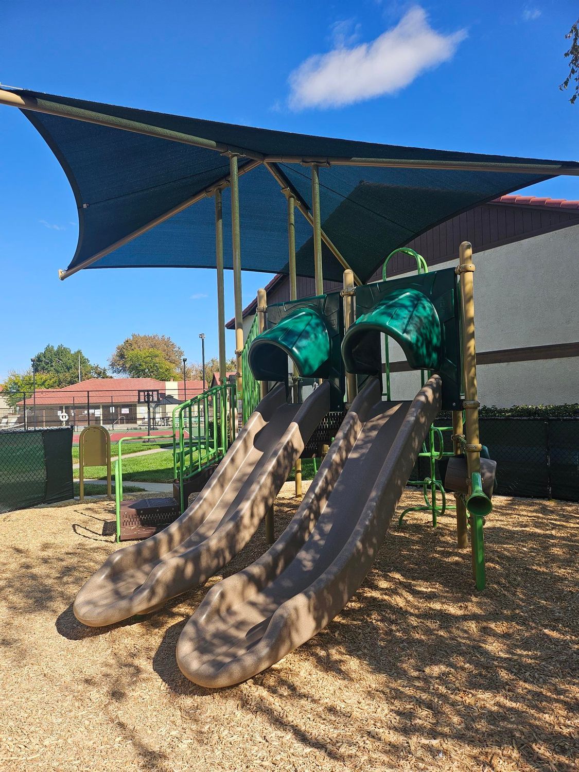 Playground with a slide, surrounded by sand, near an apartment building under a blue sky.