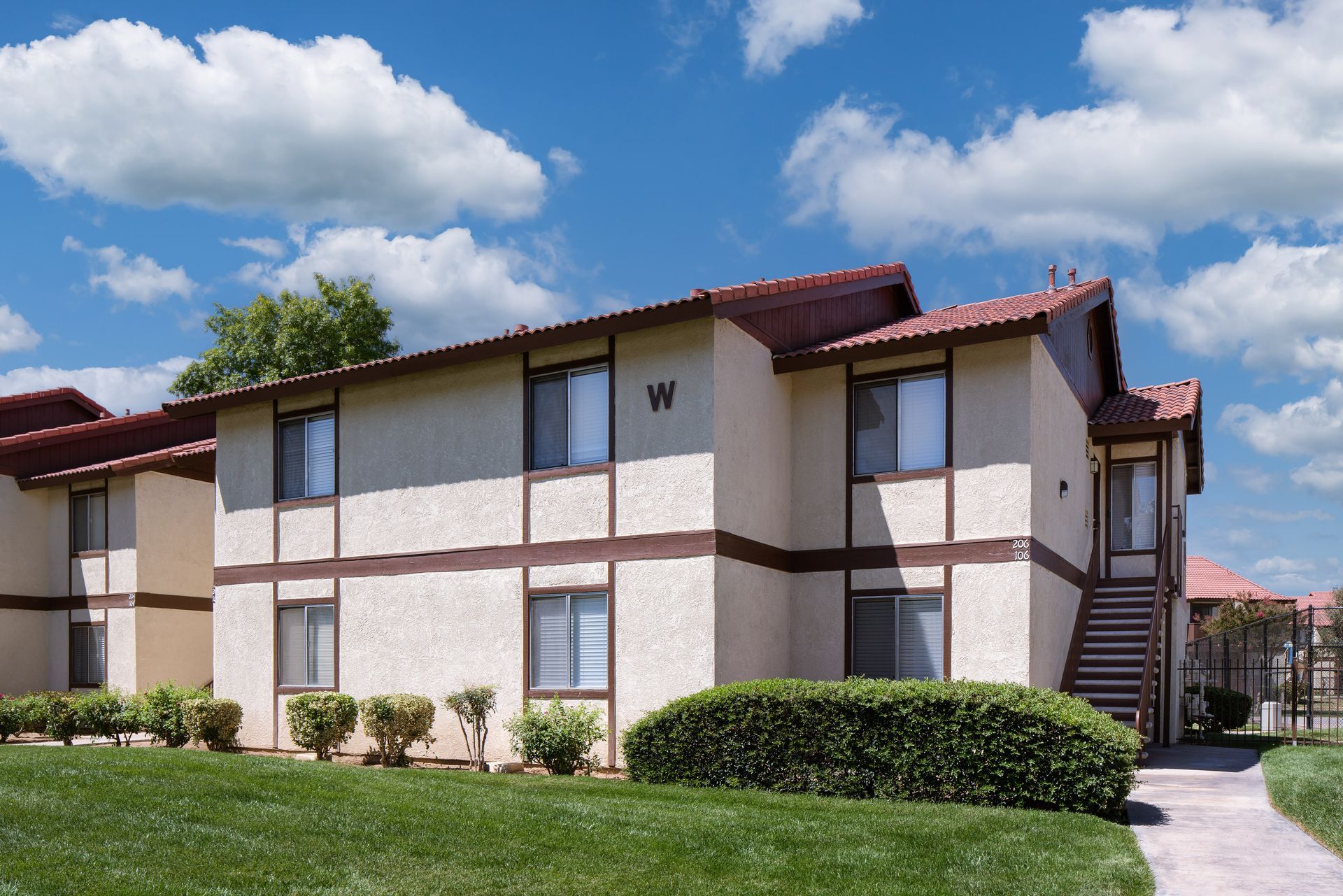 Apartment complex exterior; tan stucco walls, brown trim, red tile roof, green lawn, blue sky with clouds.