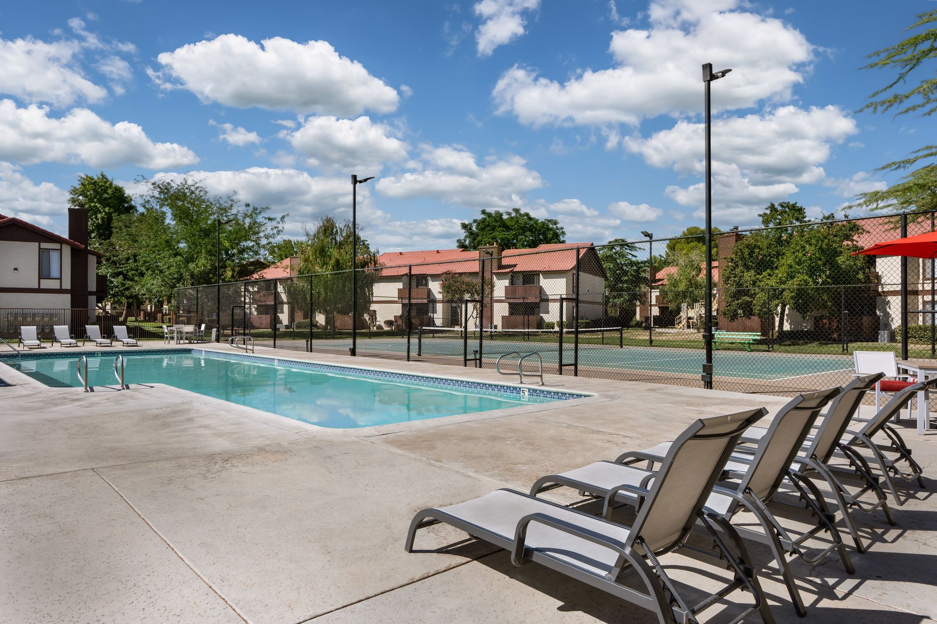 Pool area with lounge chairs, a clear pool, and apartment buildings under a blue sky with fluffy clouds.