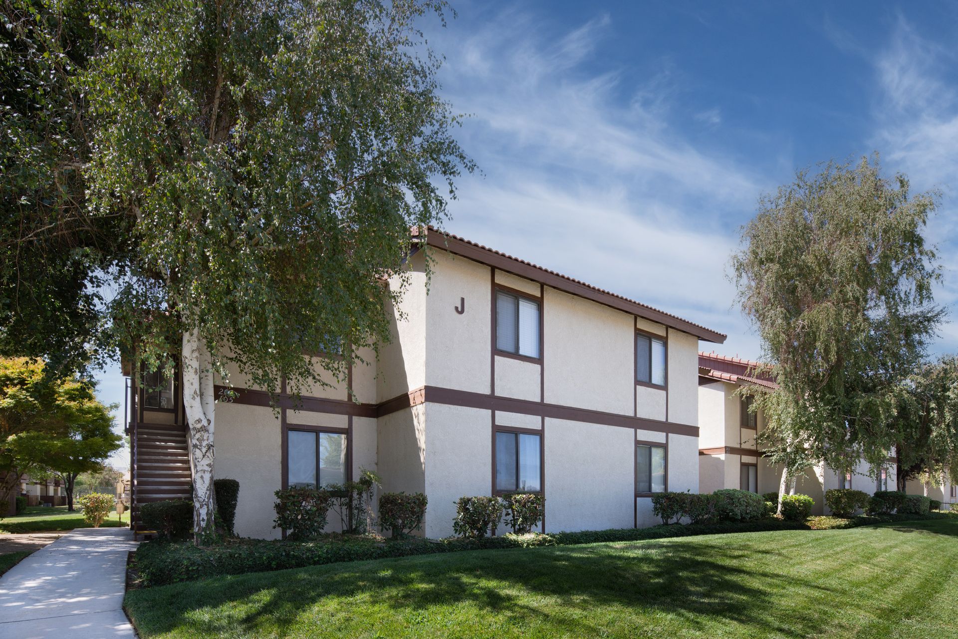 Two-story beige apartment building with brown trim, green lawn, trees, and blue sky.