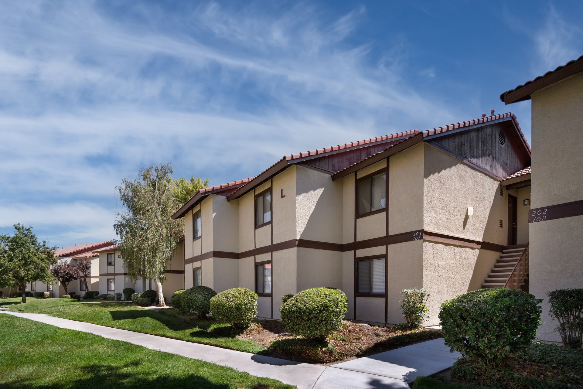 Apartment complex with beige buildings, red tile roofs, and green lawns under a blue sky.