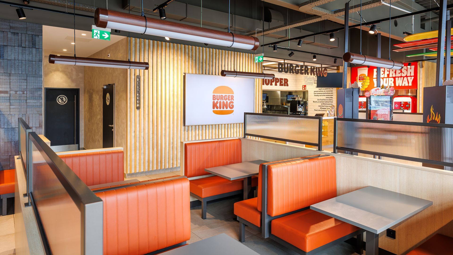 Restaurantumbau Burger King Vösendorf - Moderner Sitzbereich im Gastronomie-Interieur mit orangefarbenen Polsterbänken, Holzelementen an Wand und Tischen und einem großen Digital-Display  – Referenzprojekt Ladenbau und Innenausbau Generalunternehmer