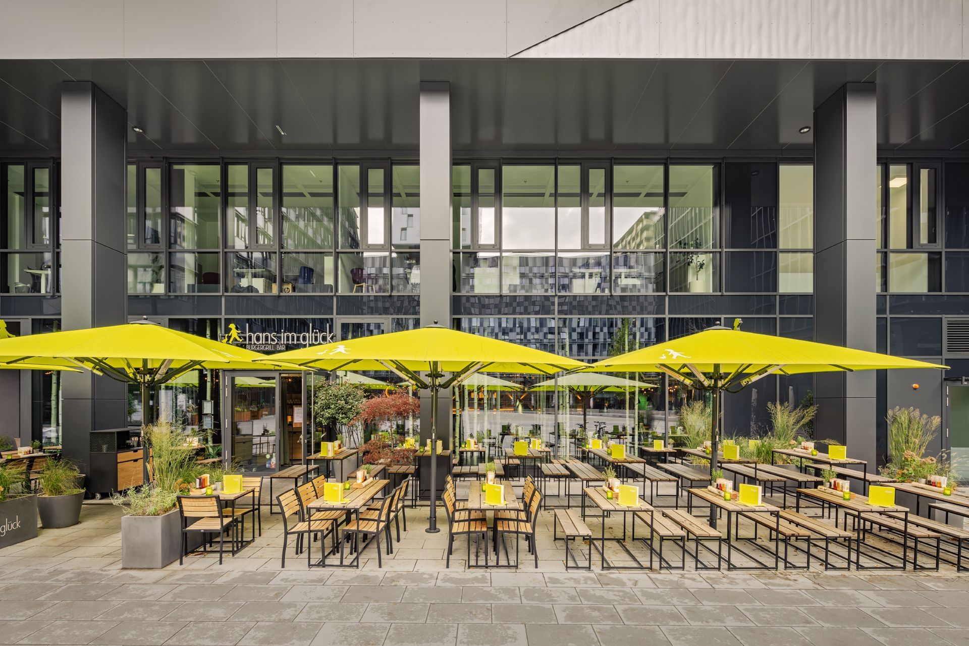 Hans im Glück Wien - Moderner Restaurant Außenbereich mit grünen Sonnenschirmen, maßgeschneiderten Möbeln, vor modernem Glasgebäude – Referenzprojekt Restaurantumbau Generalunternehmer Innenausbau