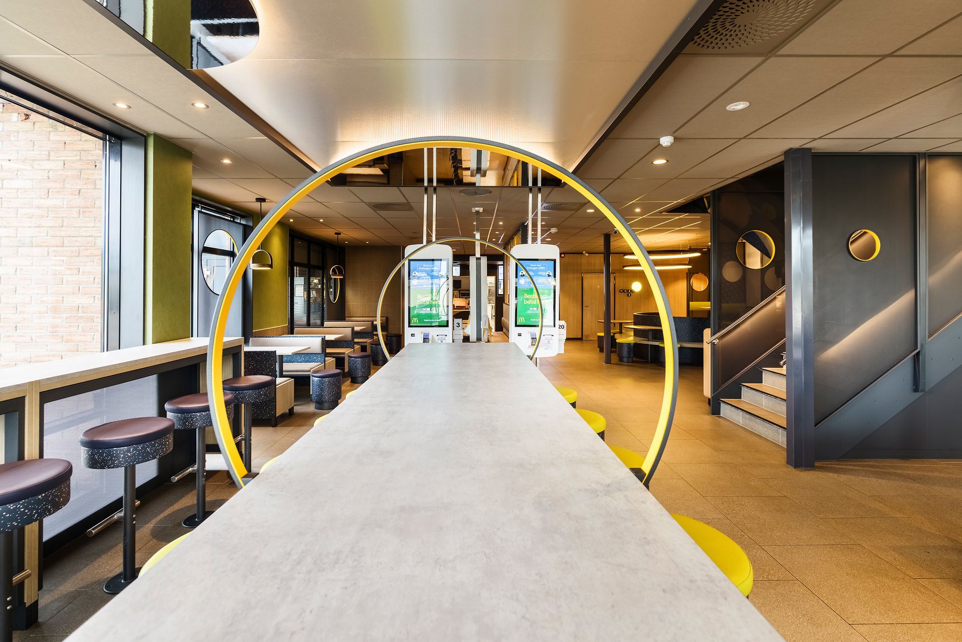 McDonald's Luna Gardermoen - modernen Fast-Food-Restaurant, Sitzgelegenheit und bogenförmige LED-Lichtelement in kräftigem Gelb über Tisch mit Betonoptik - Referenzprojekt Generalunternehmer Innenausbau