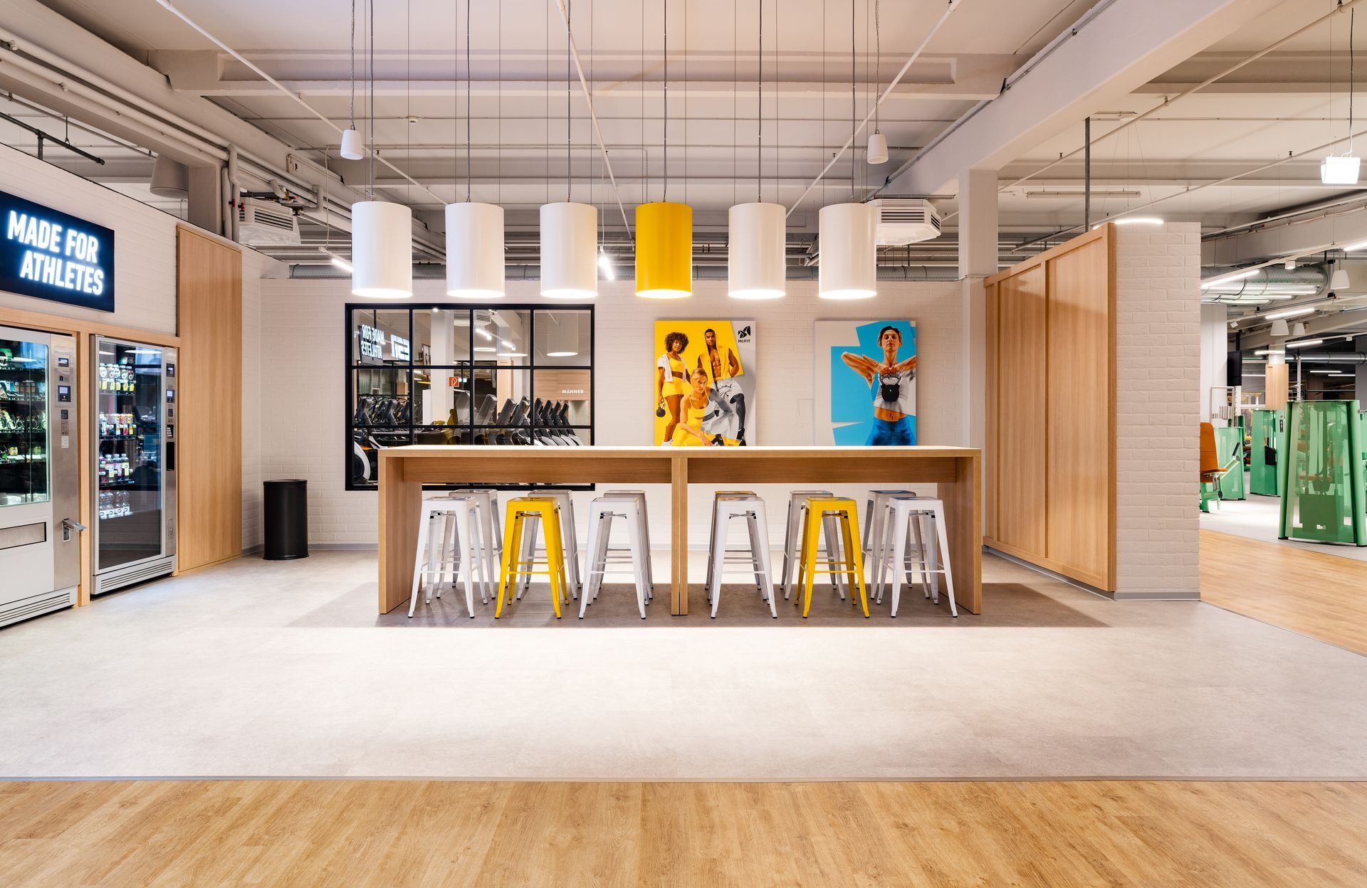 Innenausbau Fitnessstudio McFIT München Laim - Helle, moderne Lounge im Innenausbau mit hoher Bartheke, gelben Hockern, stylischen Deckenlampen und farbenfrohen Wandbildern - Referenzprojekt Fitnessstudio Innenausbau Generalunternehmer