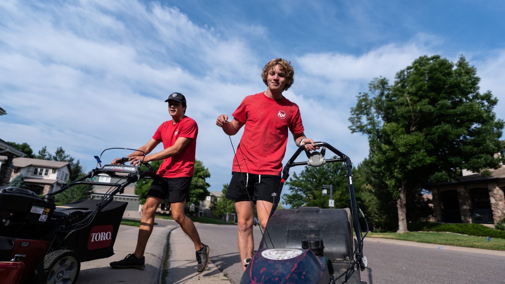 Two men are pushing a lawn mower down the sidewalk.