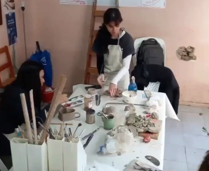 Personas trabajando en una mesa con arcilla y herramientas en un taller. Una de ellas lleva delantal.