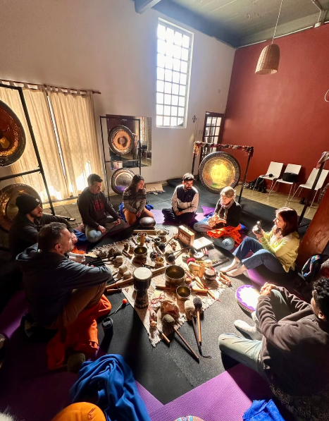 Personas sentadas en círculo alrededor de instrumentos y gongs. Ambiente interior con luz natural.