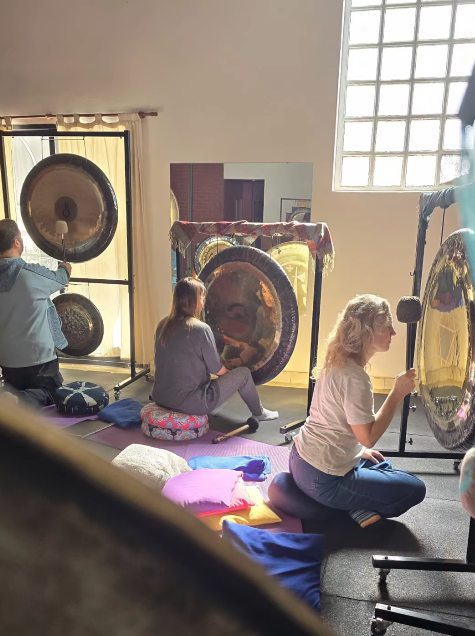 Personas tocando gongs en una sala, sentadas sobre cojines. Gongs dorados de varios tamaños sobre soportes, con luz natural.