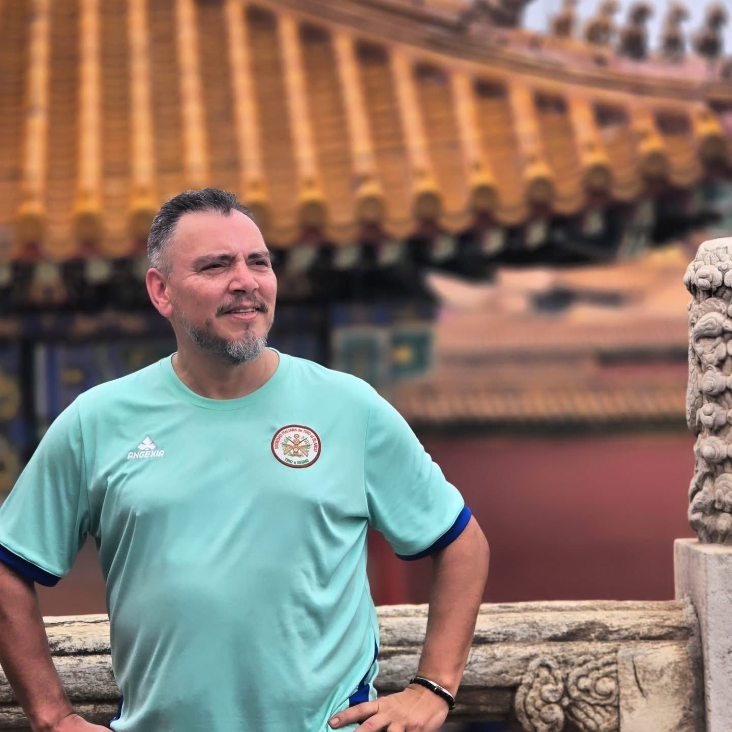 Un hombre con camisa verde azulado posa en un balcón con techo dorado adornado y tallas de piedra en China.