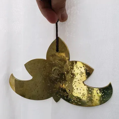 Mano sosteniendo un adorno dorado en forma de loto con un cordón, sobre un fondo blanco.