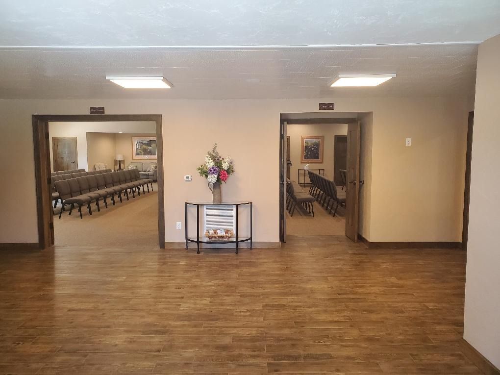 Empty lobby with wood floors, beige walls, and open doorways leading to dining rooms, with flowers on a small table.
