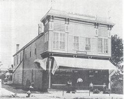 Vintage black-and-white photo of a two-story corner building with awnings and people standing outside