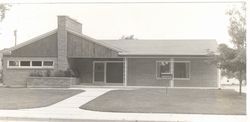 Black-and-white photo of a small brick building with a peaked roof and front entrance, viewed from the street