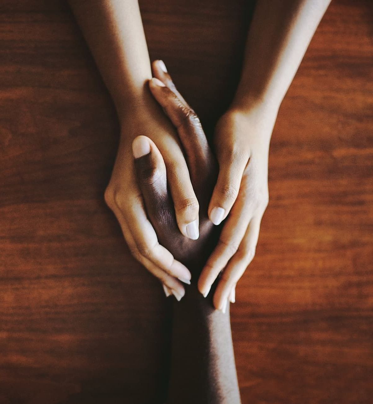 Three hands gently touching on a wooden table, lit warmly from above.