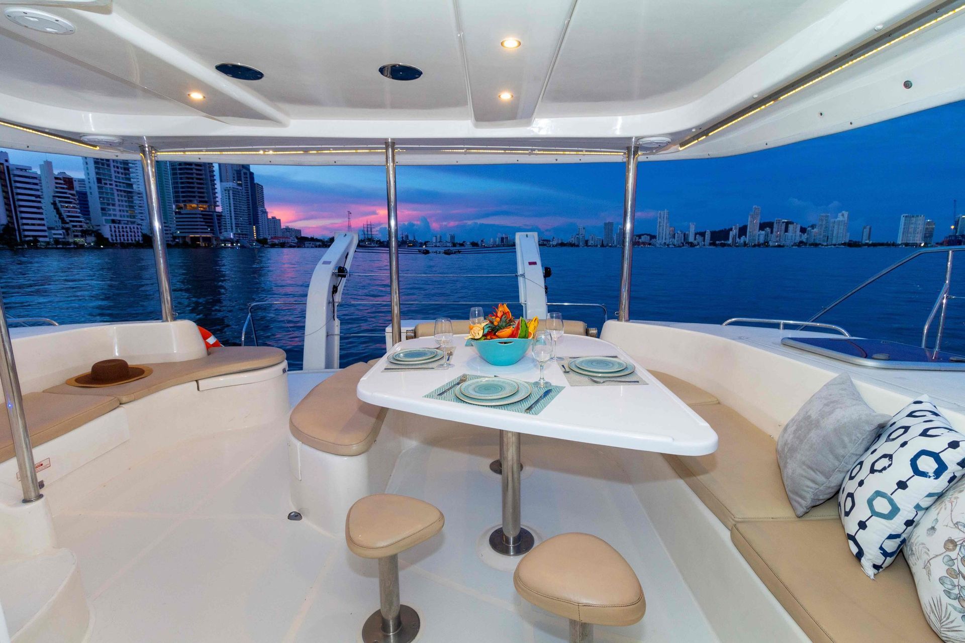 View from a yacht at dusk, overlooking a city skyline. A dining table with place settings is set up, surrounded by seating.