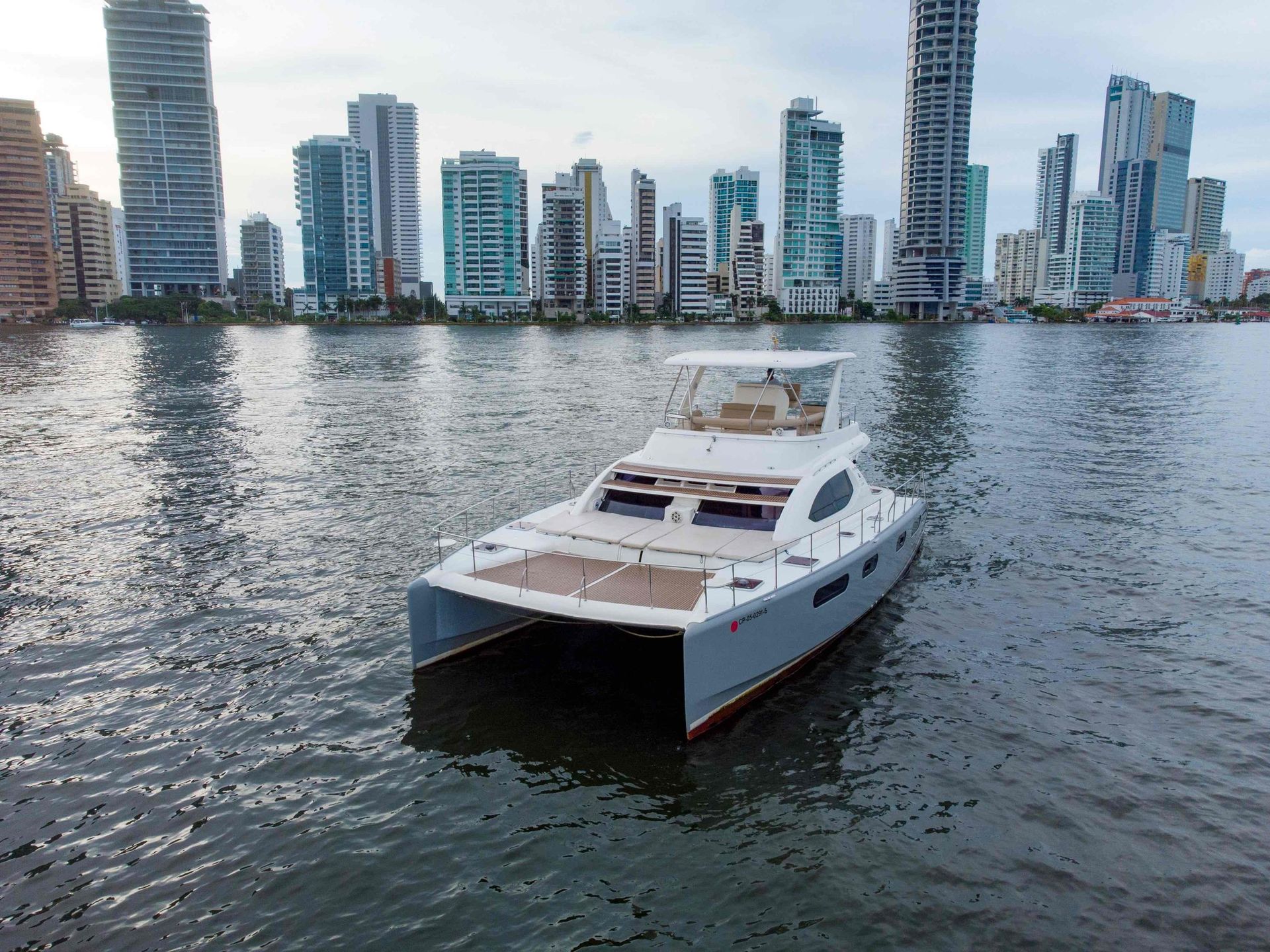 Catamaran on water with a city skyline in the background; gray and white boat, clear water, and tall buildings.