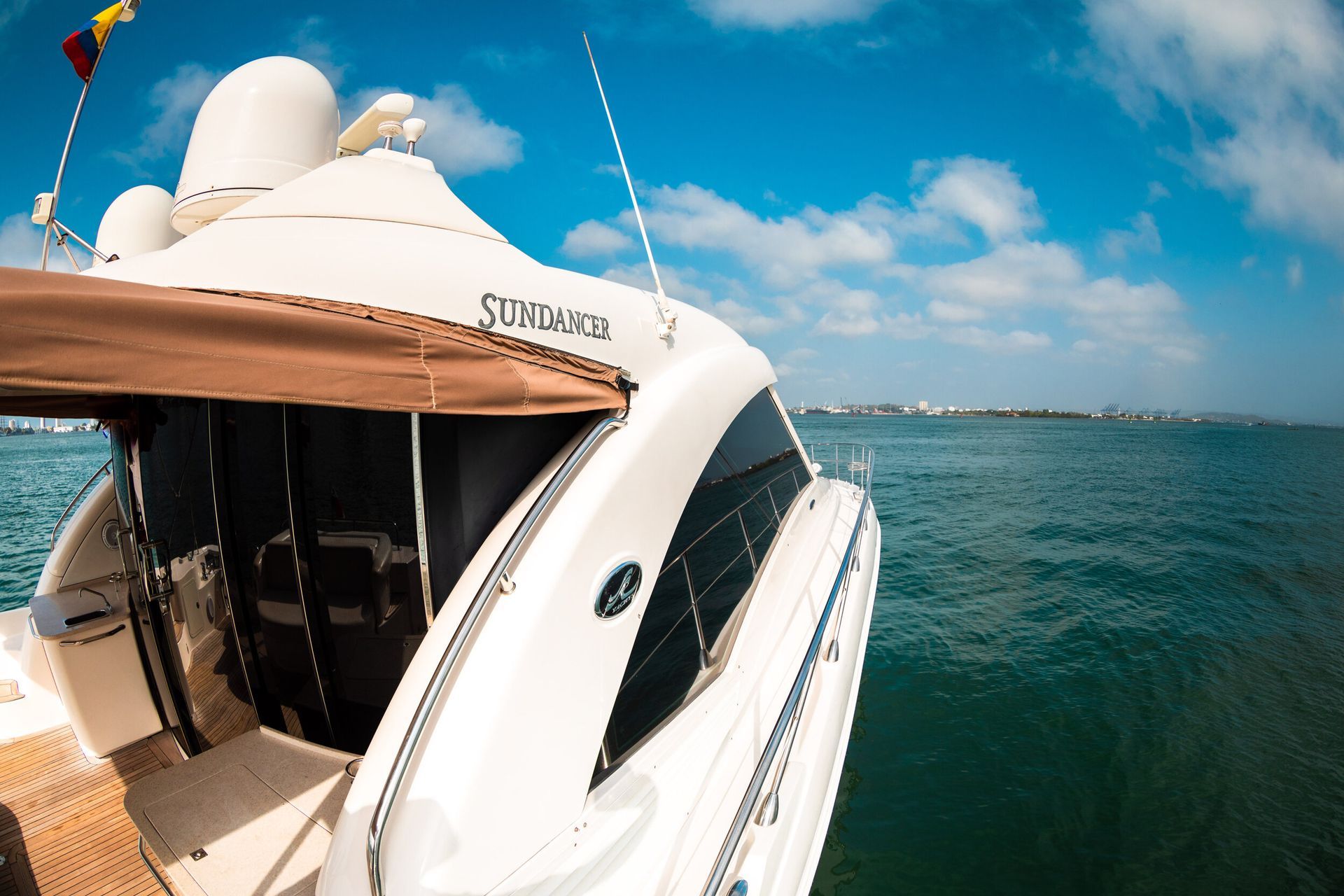 A white yacht with antennas on the water, blue sky, and a glimpse of a coastal city in the distance.
