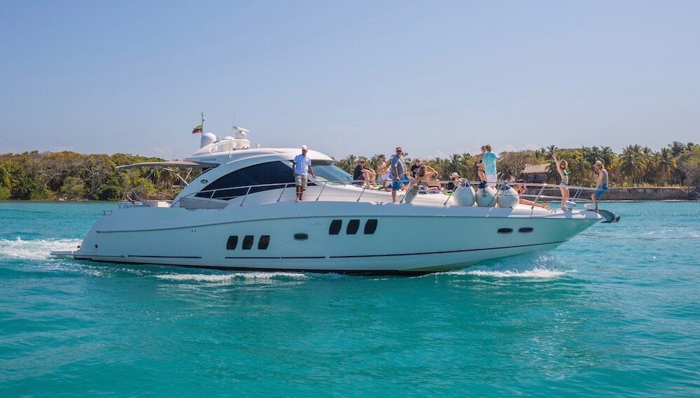 White yacht sailing on turquoise water with people on deck near a tropical shoreline.