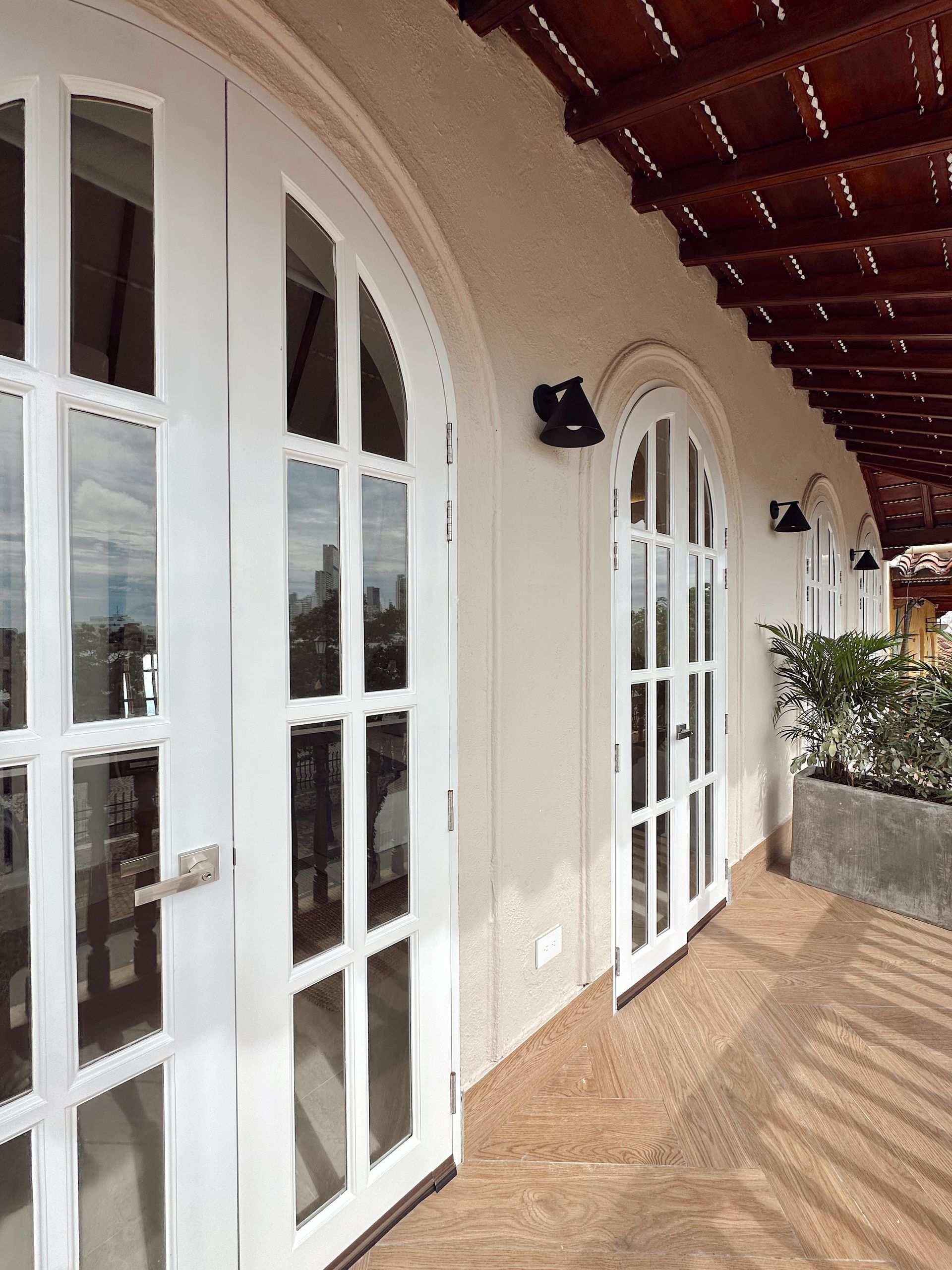 White arched doors open onto a balcony with brown flooring. A wooden pergola provides shade, and a cityscape is visible in the distance.