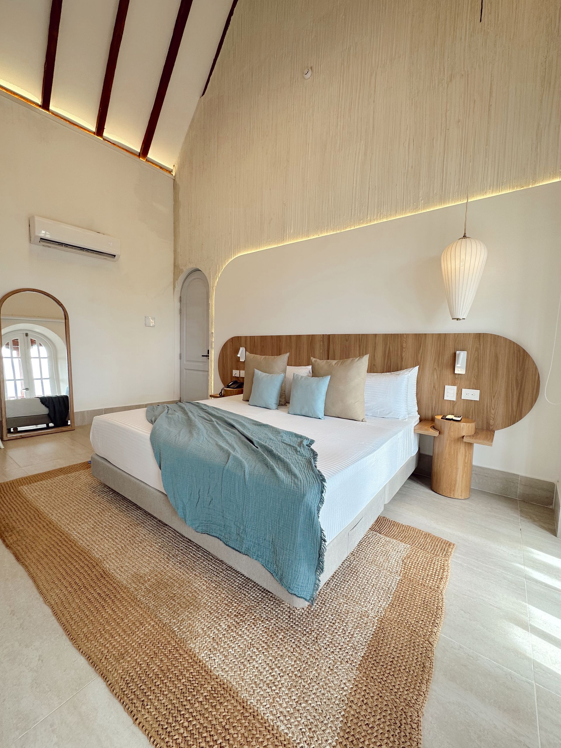 Spacious, light-filled bedroom with a king-sized bed, wooden headboard, and a rug. The room has neutral tones, a high ceiling and a large mirror.