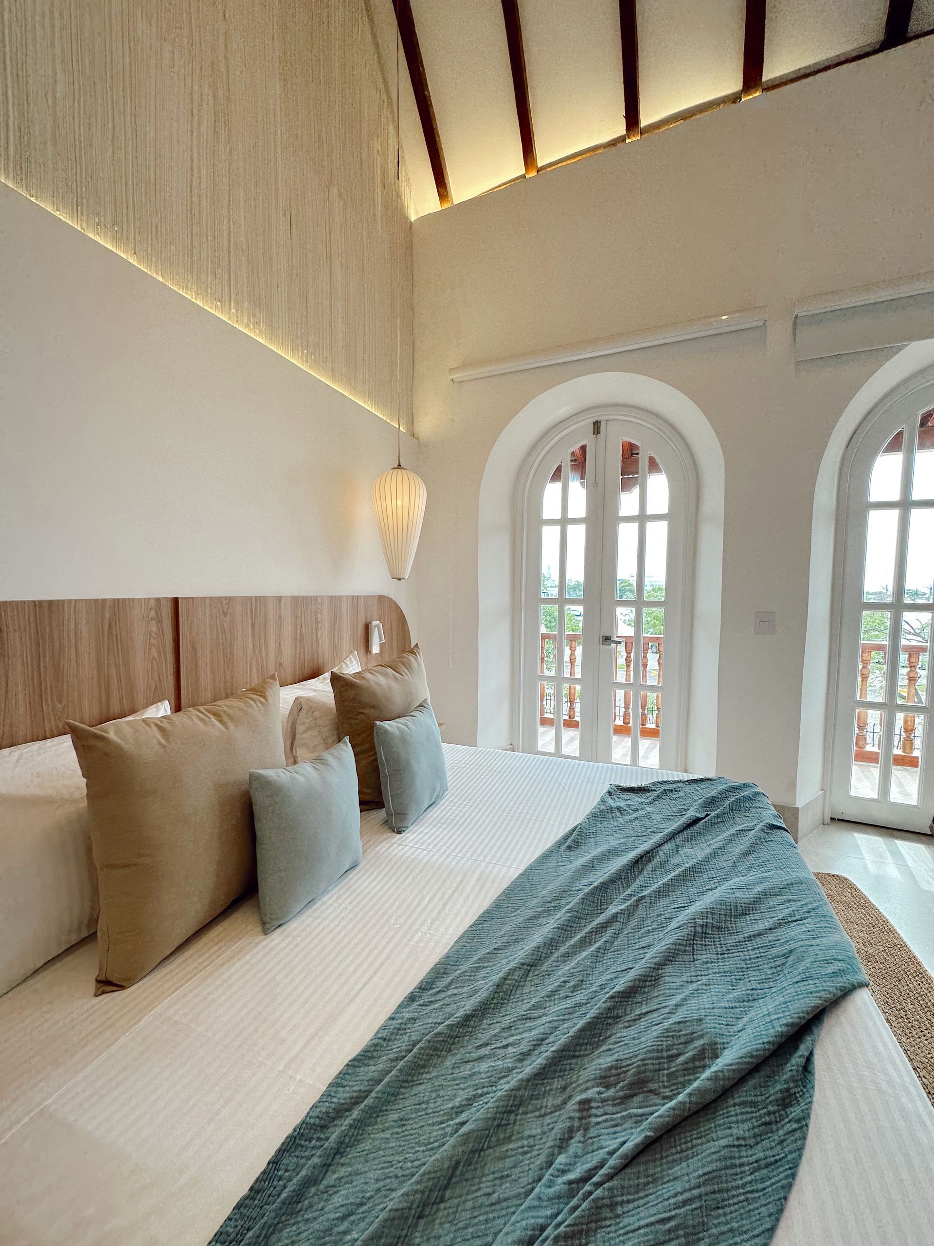 Bright bedroom with a large bed, light wood headboard, and blue quilt. White arched doors and windows lead to a balcony.