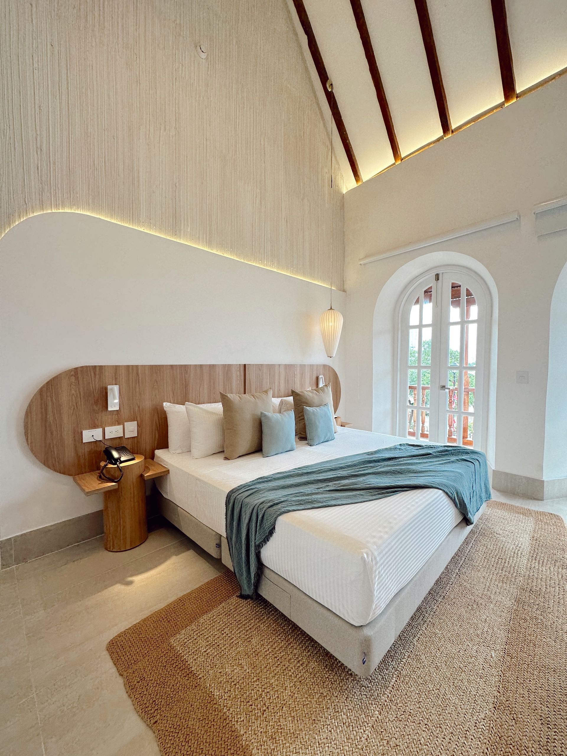 Bedroom with a large bed, wooden headboard, and woven rug. The room is bright with white walls and an arched doorway leading to a balcony.