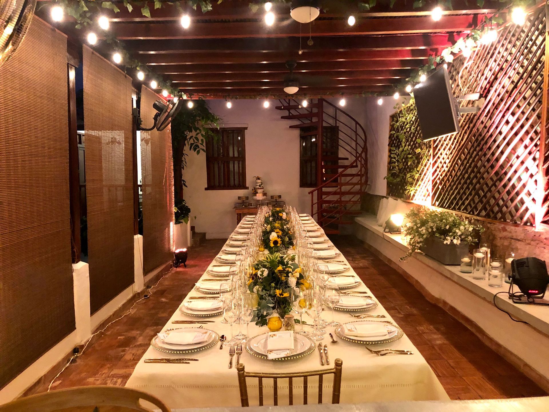 Long table set for a celebration, outdoors under string lights. Floral centerpieces adorn the table in a brick-paved patio setting.