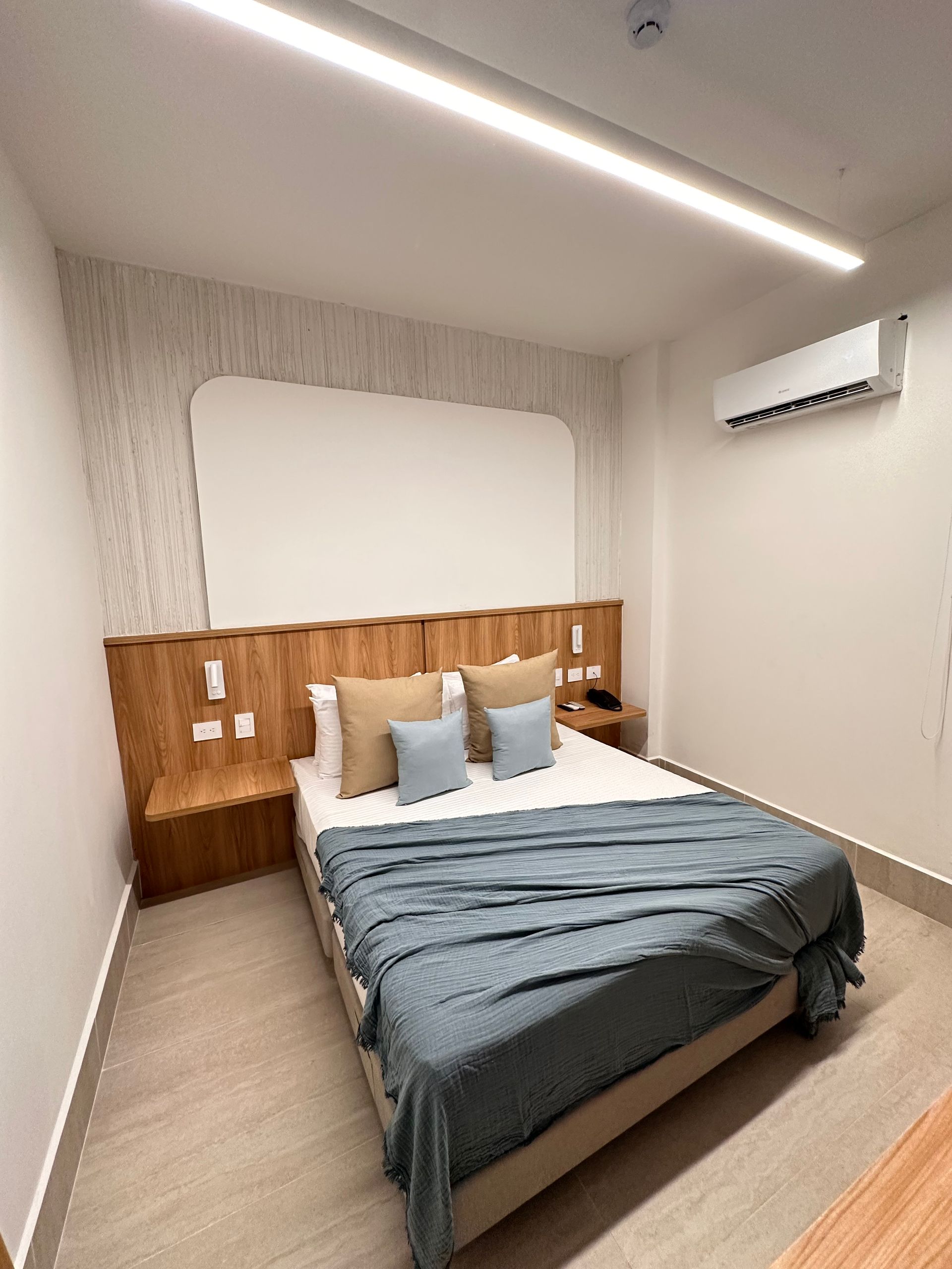 A modern hotel bedroom with a bed, wooden headboard, and air conditioner. The bed has blue bedding and tan pillows.