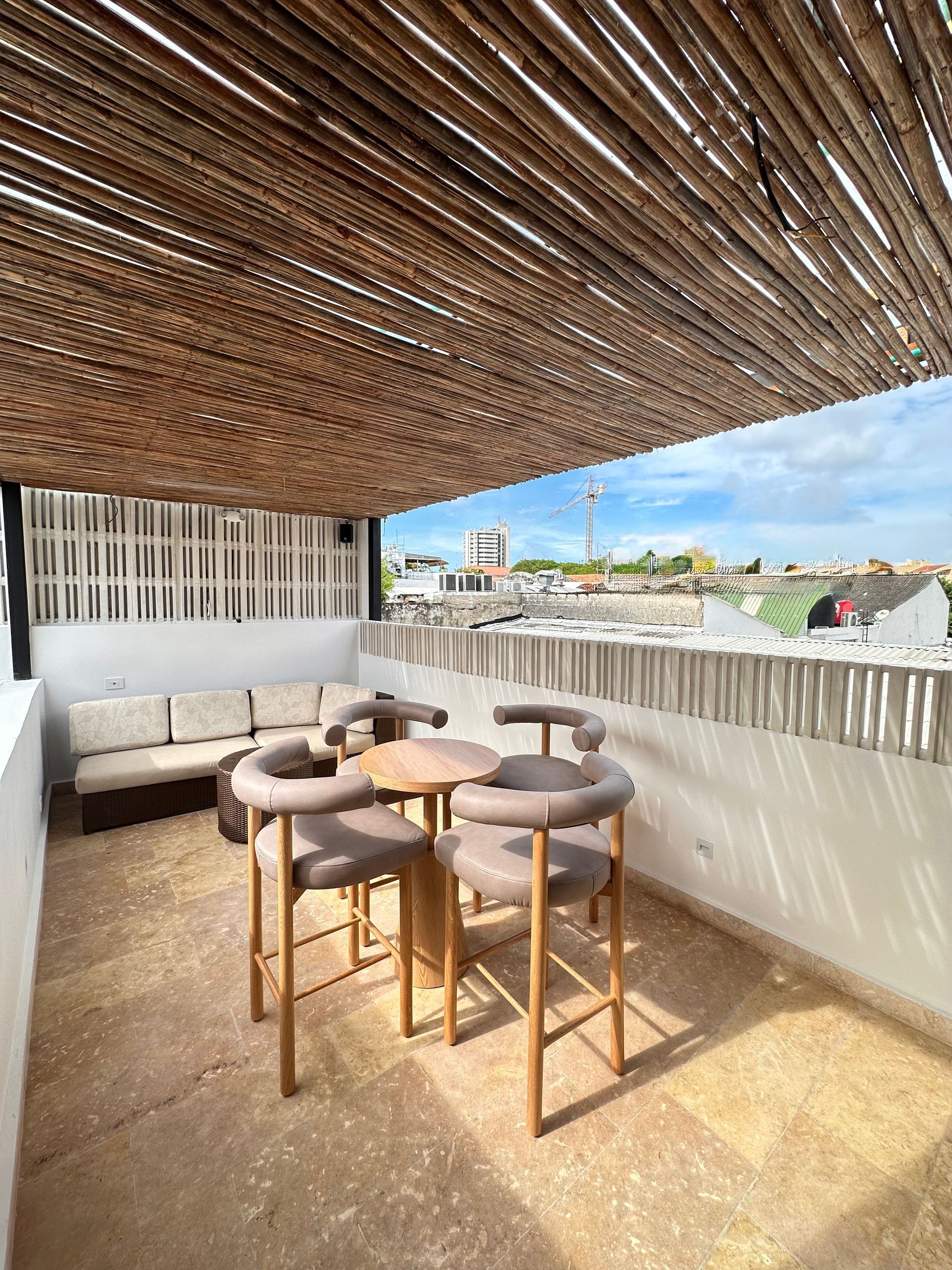 Rooftop patio with a woven reed ceiling, seating area with chairs around a small table, and a sofa against a white wall.