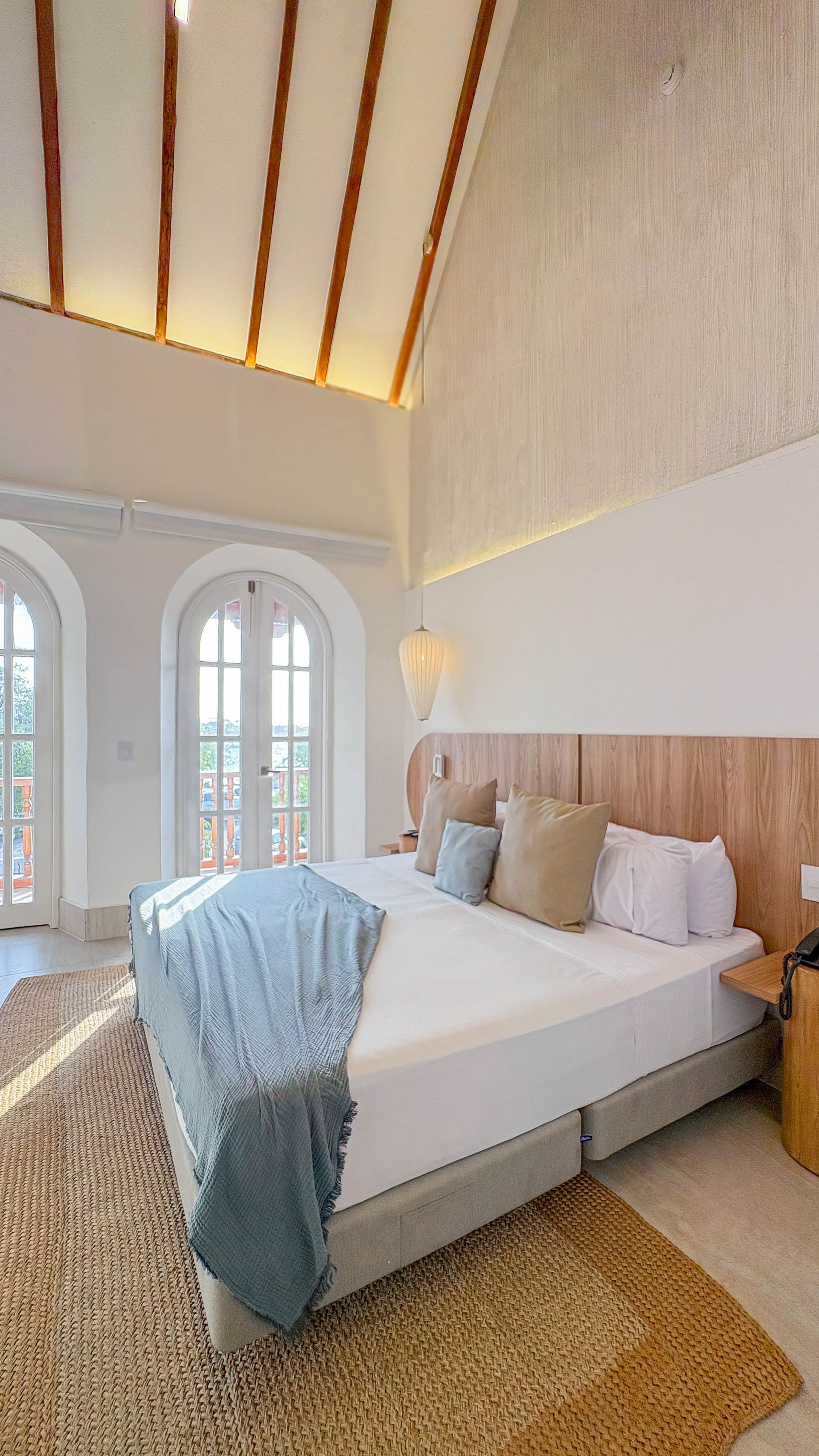 Bedroom with a high, vaulted ceiling, a bed with light-colored linens, and arched doors leading to a balcony. The room has light walls and a woven rug.