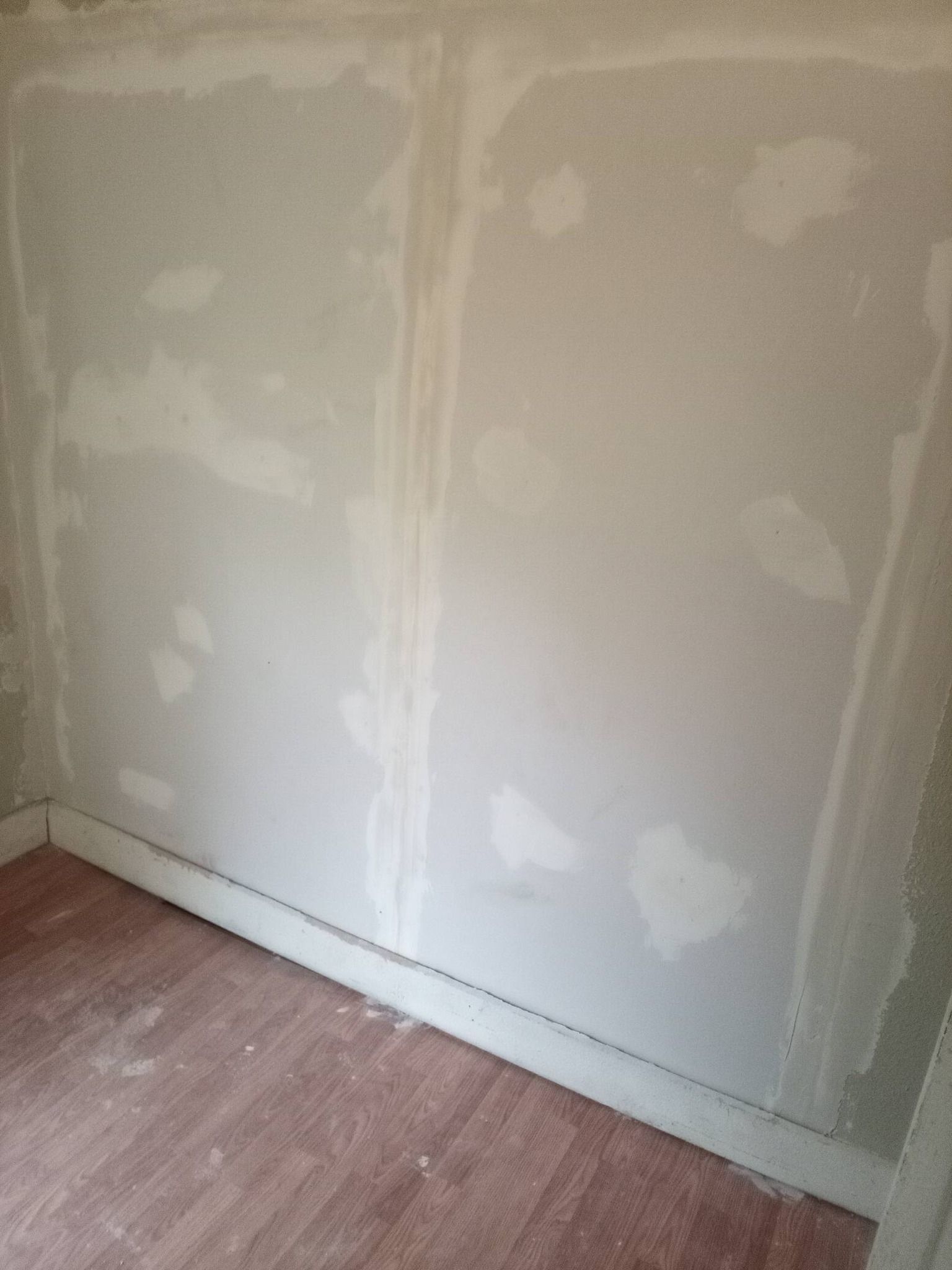Unfinished drywall wall with white joint patches above a wood floor and baseboard trim.