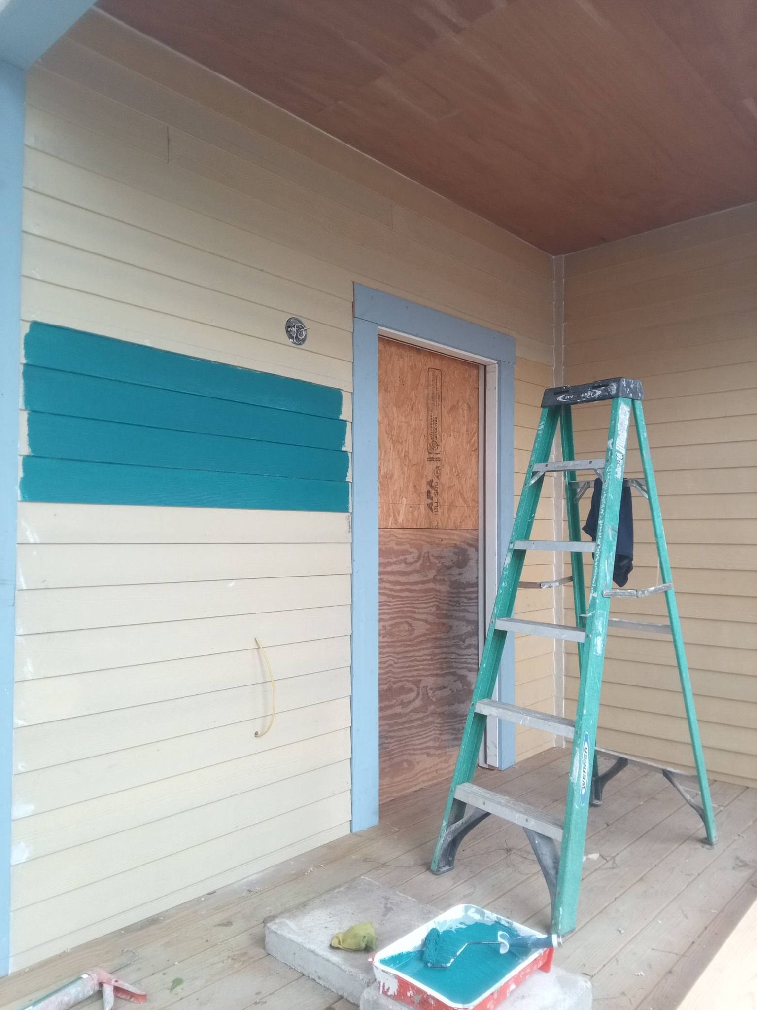 A porch wall being painted cream and teal, with a ladder and paint tray in front of a screened door.