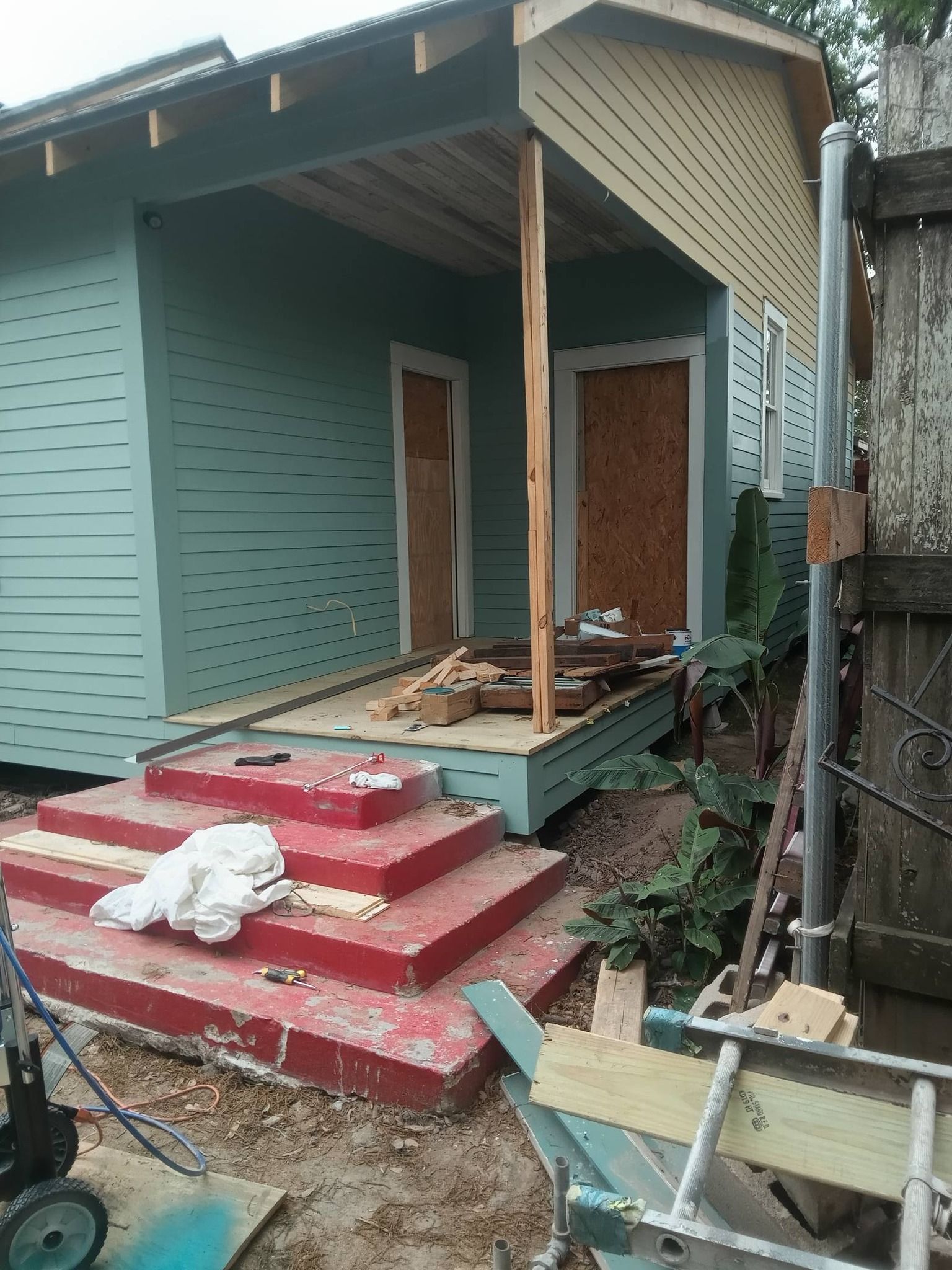 Damaged front porch of a teal house with broken steps, missing railings, and scattered debris