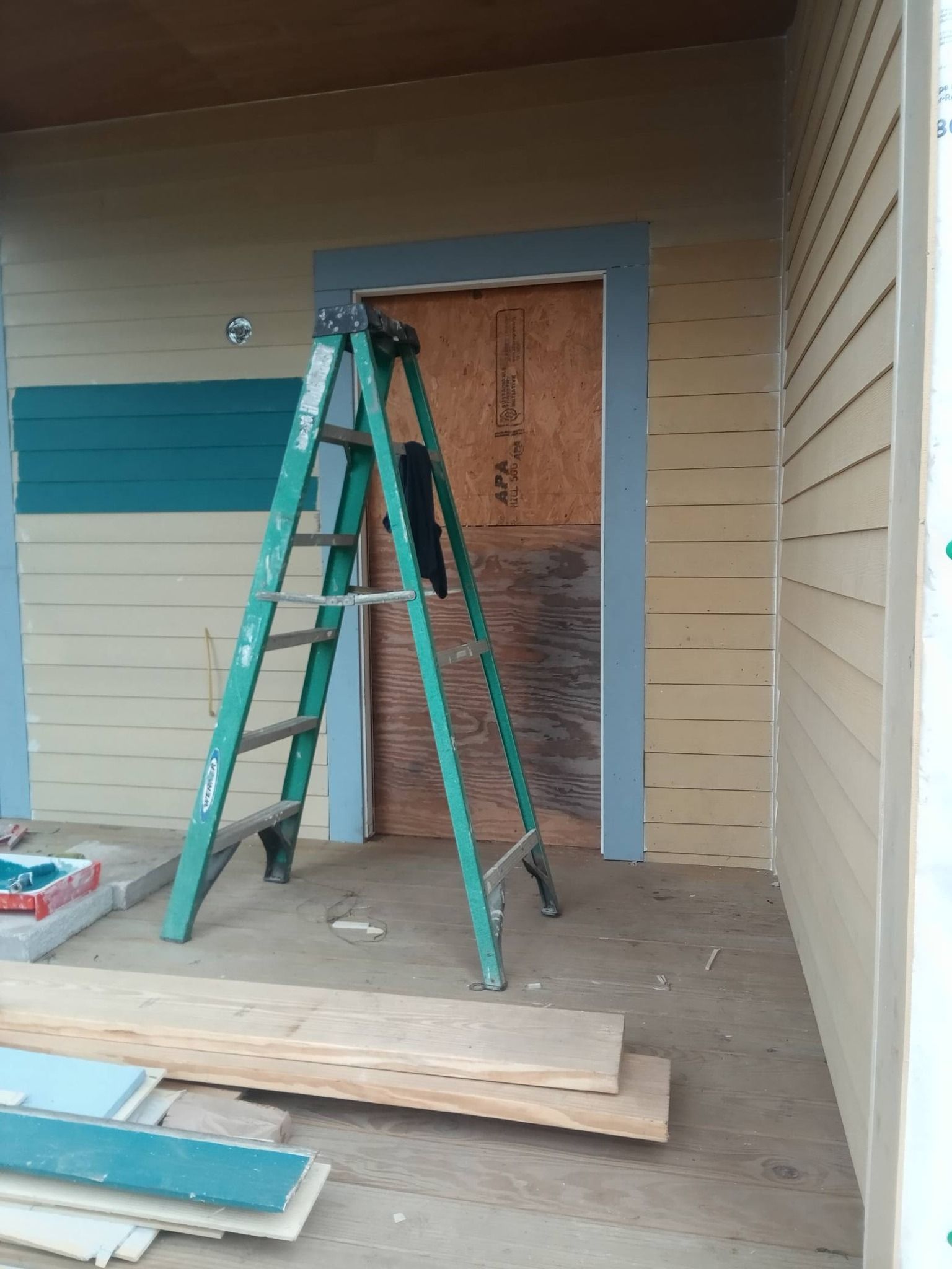 Green ladder on a porch under renovation, beside a boarded doorway and stacked lumber.
