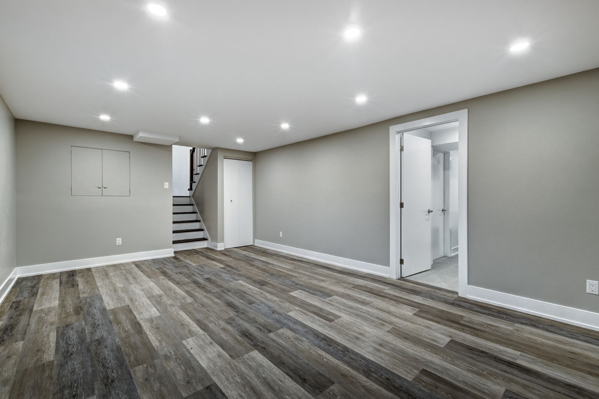 Empty modern basement room with gray walls, wood-look flooring, recessed lights, stairs, and a doorway.