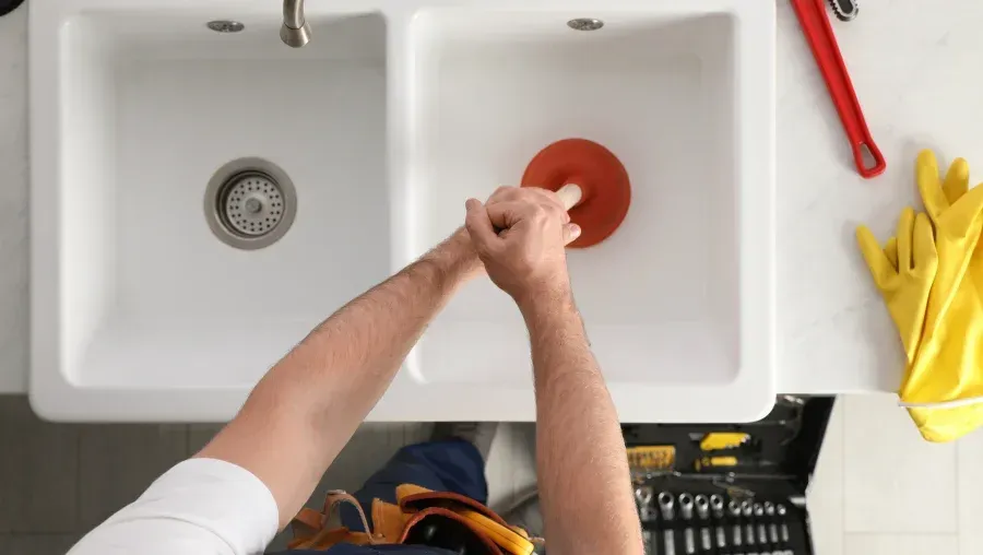 Person using a red sink plunger in a white double kitchen sink, with yellow rubber gloves nearby