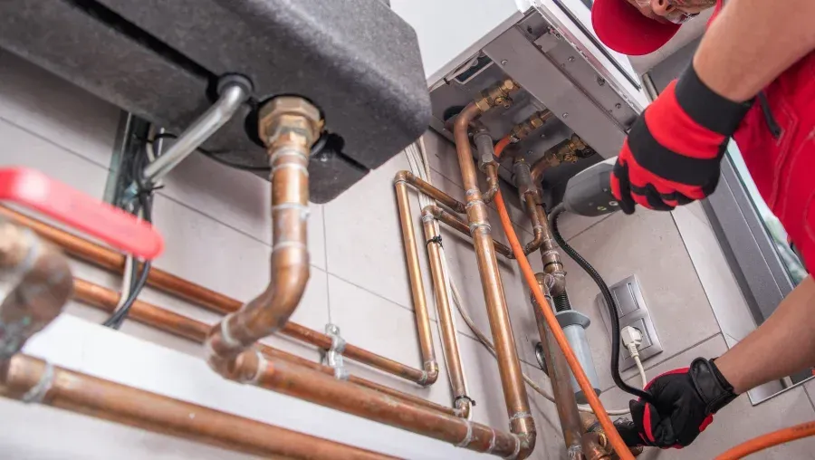 Plumber repairing copper pipes under a sink, wearing red gloves and using tools in a tight utility space