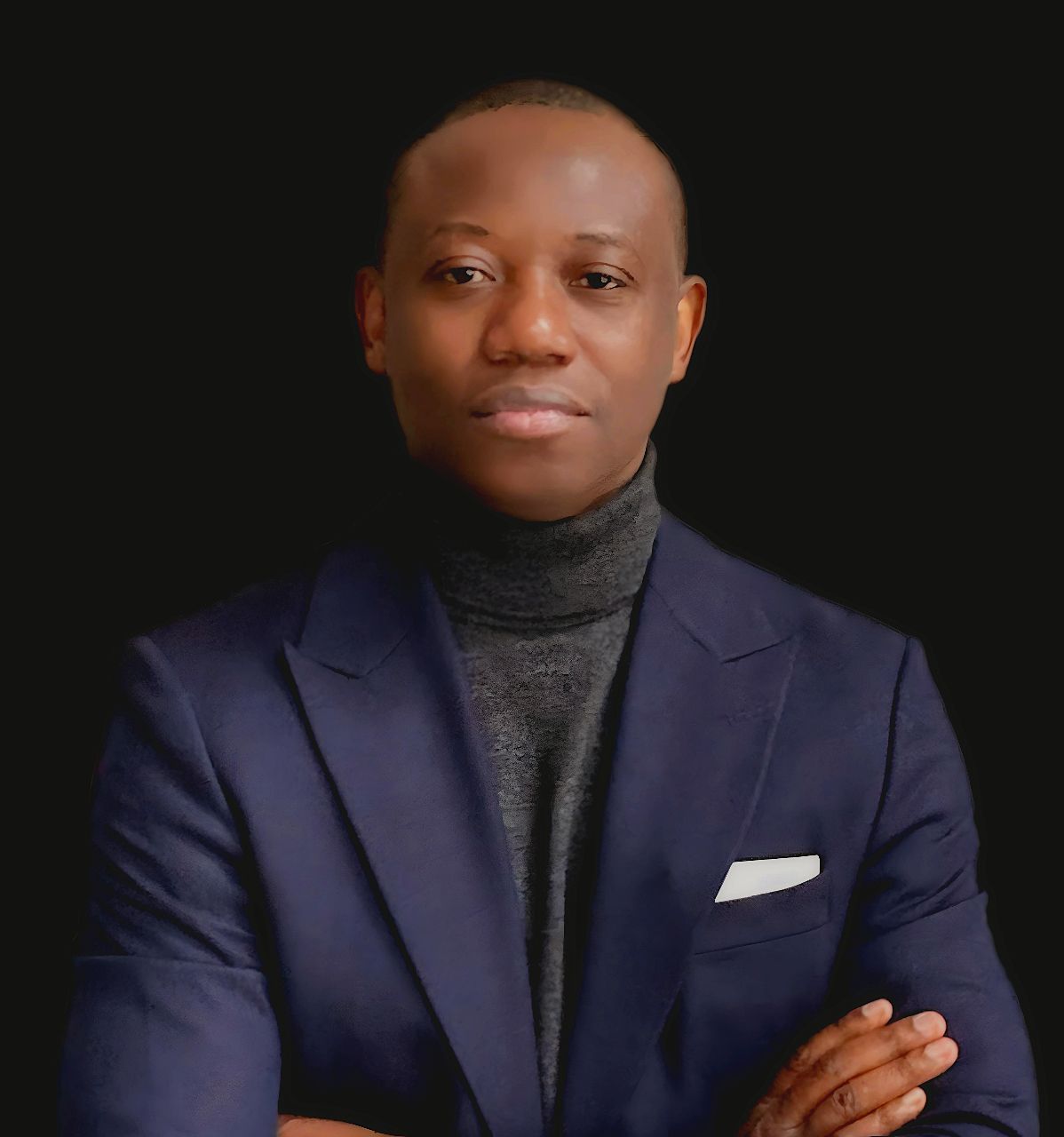 Studio portrait of a person in a navy blazer and dark turtleneck, arms crossed, against a black background