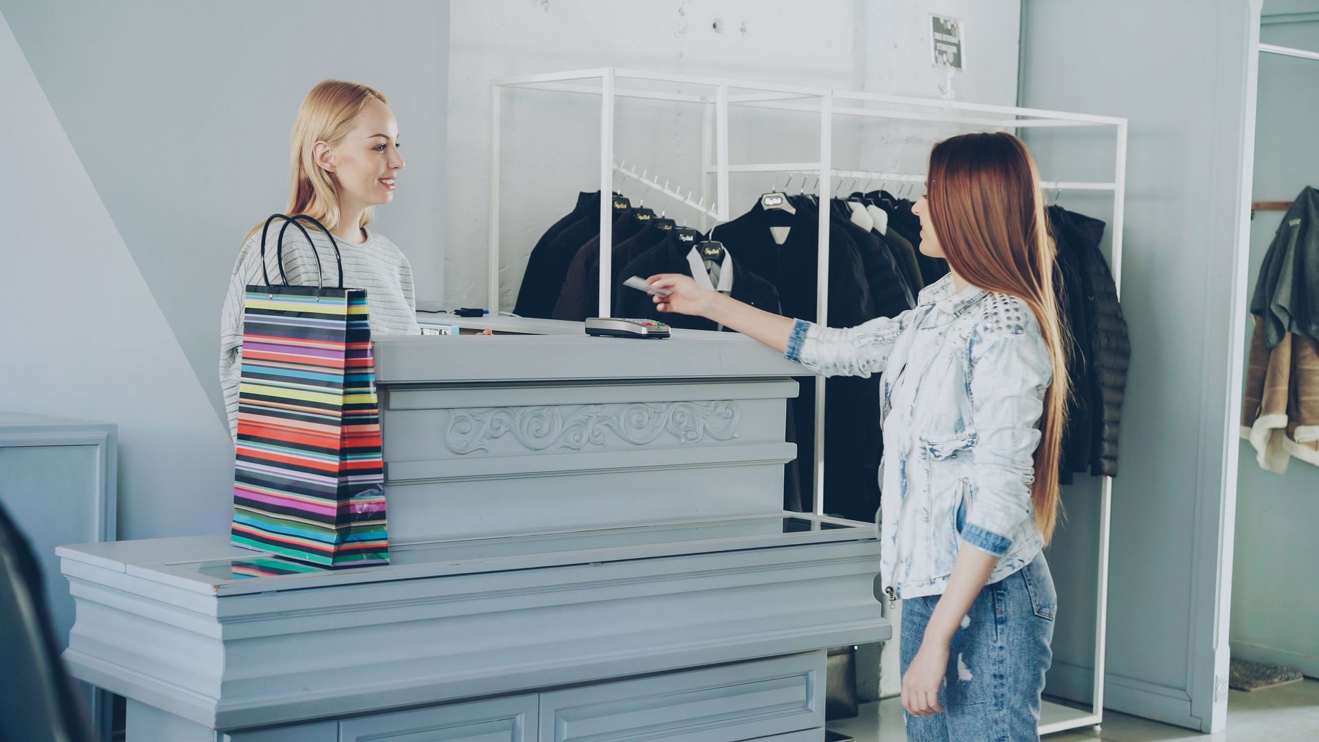 Two people in a clothing store, one holding a striped shopping bag and one pointing at folded clothes.