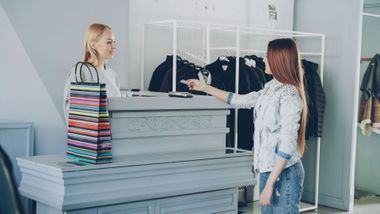 Two people in a clothing store, one holding a striped shopping bag and one pointing at folded clothes.