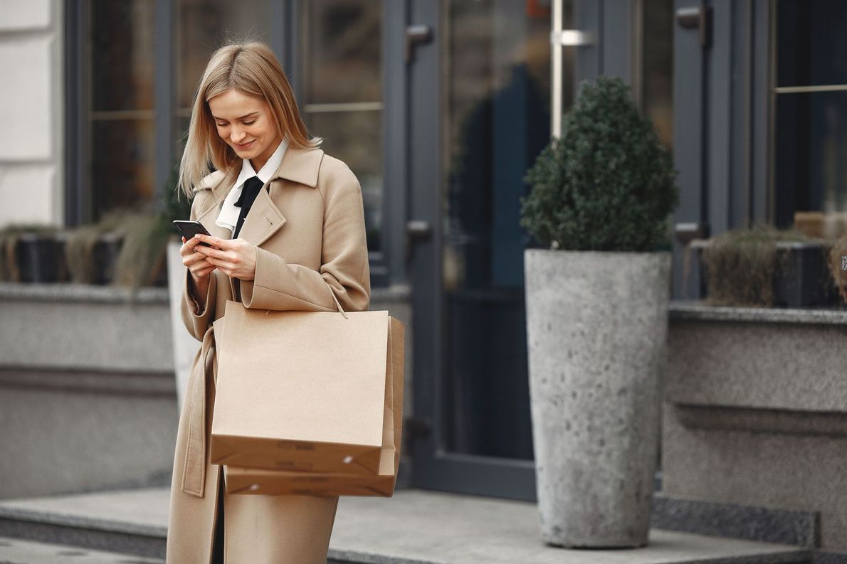 Woman in a beige coat using a smartphone while walking with shopping bags outside a store.